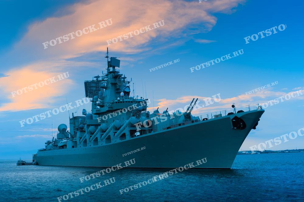 Военный корабль стоит на якоре в открытом море при закате. Оранжево-голубое небо создаёт впечатляющий фон, подчёркивая силуэт судна.